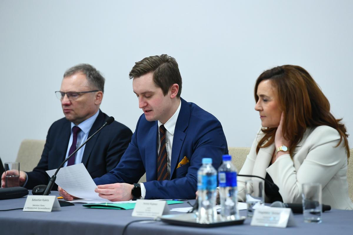 Uczestnicy konferencji, dwóch mężczyzn i kobieta siedzą przy stole prezydialnym podczas prezentacji. 