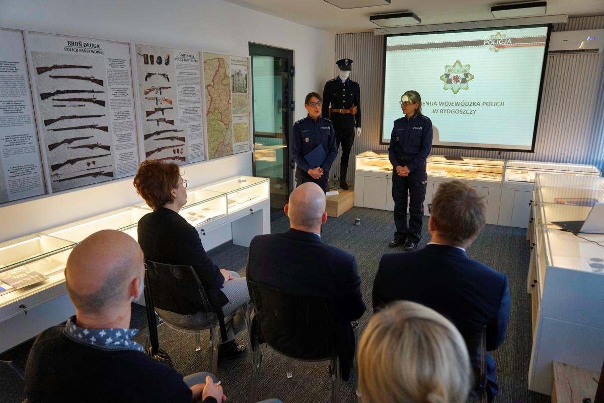 Uczestnicy siedzą na Sali konferencyjnej podczas spotkania z funkcjonariuszami Policji. Dwie kobiety w mundurach Policyjnych stoją na tle prezentacji wyświetlanej na rzutniku. 