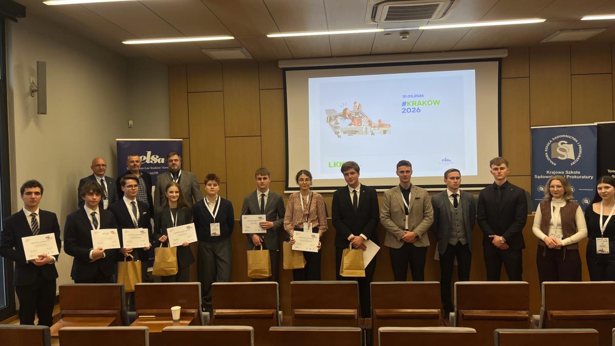 wewnątrz auli widać grupę kobiet i mężczyzn, stojących w rządzie. Osoby stojące po lewej stronie trzymają w rękach kartki oraz papierowe torebki. Za nimi, po lewej stronie stoi trzech mężczyzn. W tle na ekranie widoczny jest rysunek i napis. Z przodu na dole widoczne są dwa rzędy oparć foteli.
