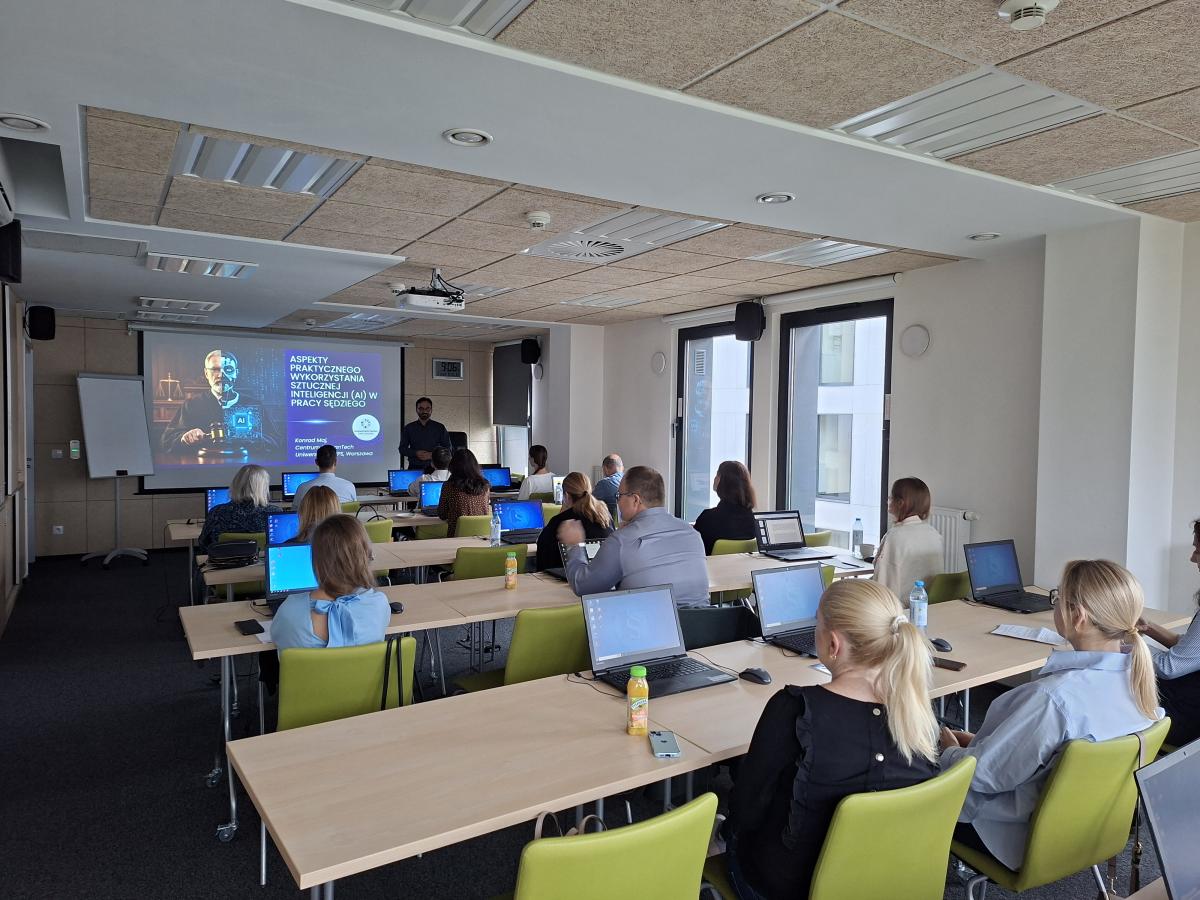 Na zdjęciu przedstawiono salę konferencyjną, wyposażoną w stanowiska komputerowe, w której odbywa się szkolenie. Uczestnicy – około 15 osób - siedzą przy długich stołach, korzystając z laptopów. Na ekranie projekcyjnym widoczny jest slajd dotyczący sztucznej inteligencji i jej zastosowań. Obok ekranu stoi wykładowca. Pomieszczenie jest jasne, nowoczesne, z dużymi oknami zapewniającymi dostęp naturalnego światła.