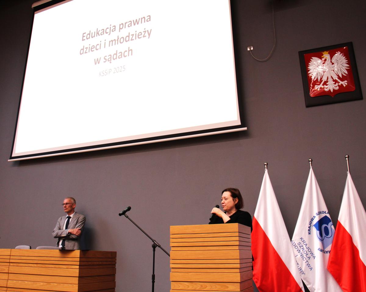 Zdjęcie przedstawia aulę z dwoma osobami stojącymi na scenie. Nad nimi znajduje się duży ekran projekcyjny z napisem „Edukacja prawna dzieci i młodzieży w sądach – KSSiP 2025”. Po prawej stronie sceny ustawione są flagi. Jedna z osób trzyma mikrofon, co sugeruje, że trwa wystąpienie lub panel dyskusyjny. 