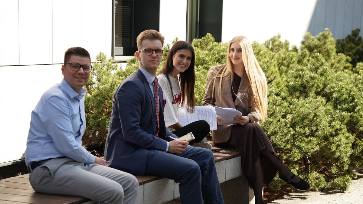 . Zdjęcie przedstawia cztery uśmiechnięte, młode osoby, w tym dwóch mężczyzn, ubrane biznesowo. Osoby te siedzą na ławce na tle ściany białego budynku i zielonych krzewów.