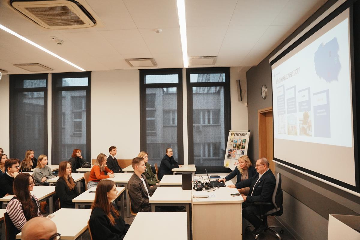 grupa około 15 osób (kobiet i mężczyzn) siedzi na sali wykładowej. Przy biurku z laptopem siedzi kobieta i mężczyzna. Za nimi widać niewyraźnie prezentację. 