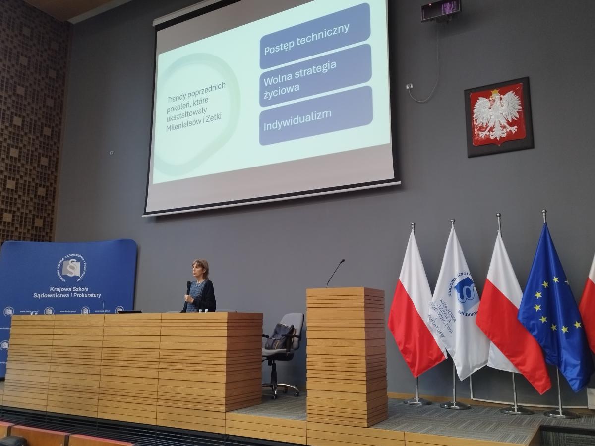 Zdjęcie przedstawia część auli wykładowej. Na pierwszym planie, za mównicą, widoczny jest mężczyzna  w ciemnym garniturze, białej koszuli i bordowym krawacie. Po prawej stronie zdjęcia znajdują się, ustawione w rzędzie, flagi: Polski, Unii Europejskiej oraz Krajowej Szkoły. W oddali, po lewej stronie, za katedrą, znajduje się baner w kolorze granatowym z napisem Krajowa Szkoła Sądownictwa i Prokuratury oraz logiem Krajowej Szkoły.