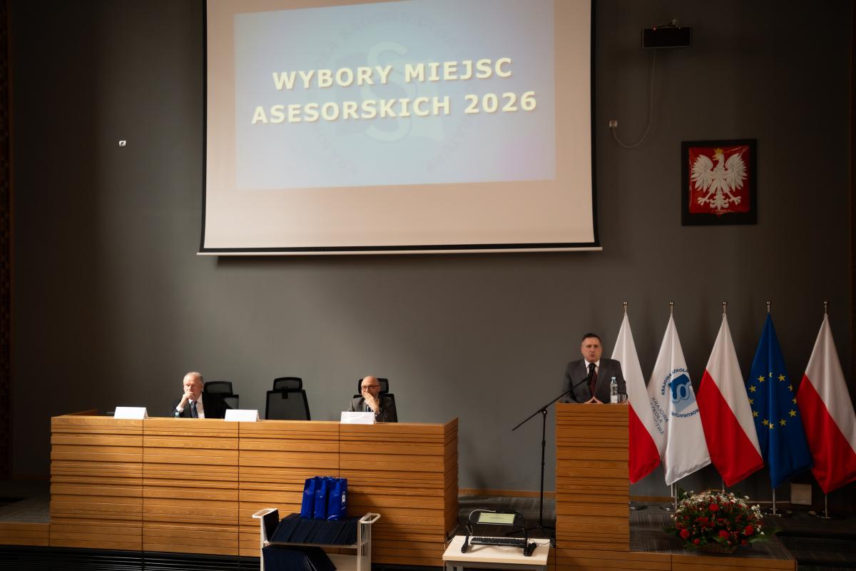 Dwóch mężczyzn siedzi przy dużym pulpicie-biurku, na którym ustawione są wizytowniki z nazwiskami. Jeden mężczyzna stoi przy mównicy, przed mikrofonem. Nad mężczyznami widoczna jest duża tablica z wyświetlonym napisem Wybór miejsc asesorskich 2026. Po prawej stronie na ścianie wisi godło Polski. Poniżej ustawione są trzy flagi Polski na zmianę z flagą Krajowej Szkoły Sądownictwa i Prokuratury oraz Unii Europejskiej.