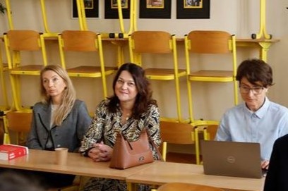 Zdjęcie przedstawia trzy osoby – kobiety, siedzące przy stole w klasie lekcyjnej. Za nimi znajduje się kilka żółtych krzeseł ustawionych na biurkach. Jedna osoba korzysta z laptopa, na stole znajduje się czerwona książka, mały kubek i torebka. Osoba pośrodku uśmiecha się.