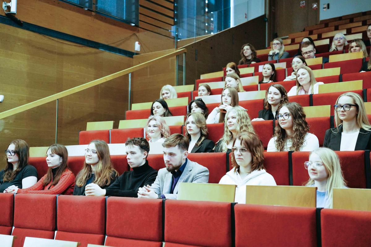 grupa ok. 30 kobiet i mężczyzn siedzi na auli wykładowej na czerwonych krzesłach.