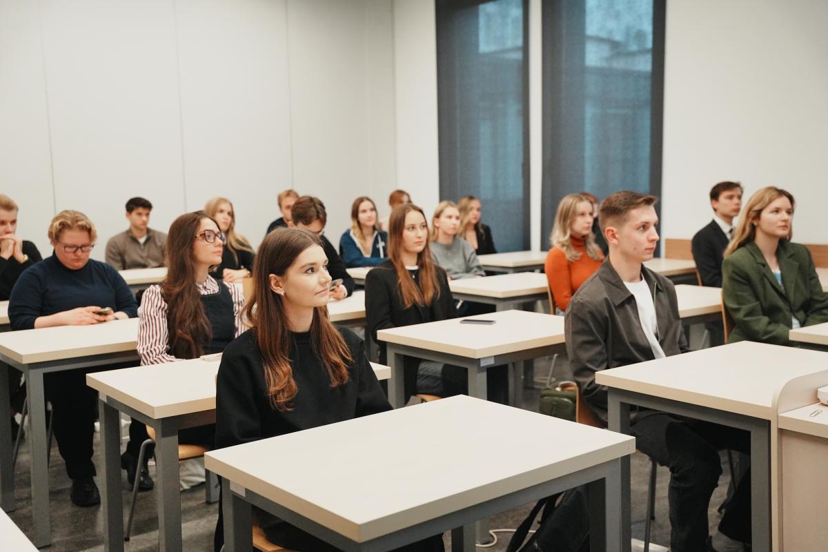 grupa około dwudziestu siedmiu kobiet i mężczyzn siedzi przy stolikach. Za nimi znajduje się duża biała ściana, a obok - po prawej stronie są duże okna zasłonięte roletami.
