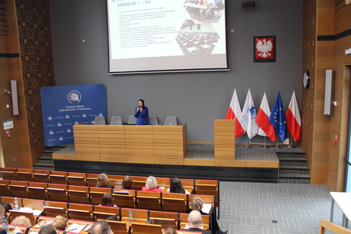 Zdjęcie przedstawia widok od środka auli wykładowej w kierunku katedry. Na zdjęciu, w jego centralnej  części, widoczna jest kobieta o ciemnych długich włosach, ubrana w kobaltowy garnitur. Kobieta stoi za katedrą, trzyma w dłoni mikrofon. Za kobietą widoczny jest, na tle szarej ściany, ekran z wyświetlaną prezentacją. 