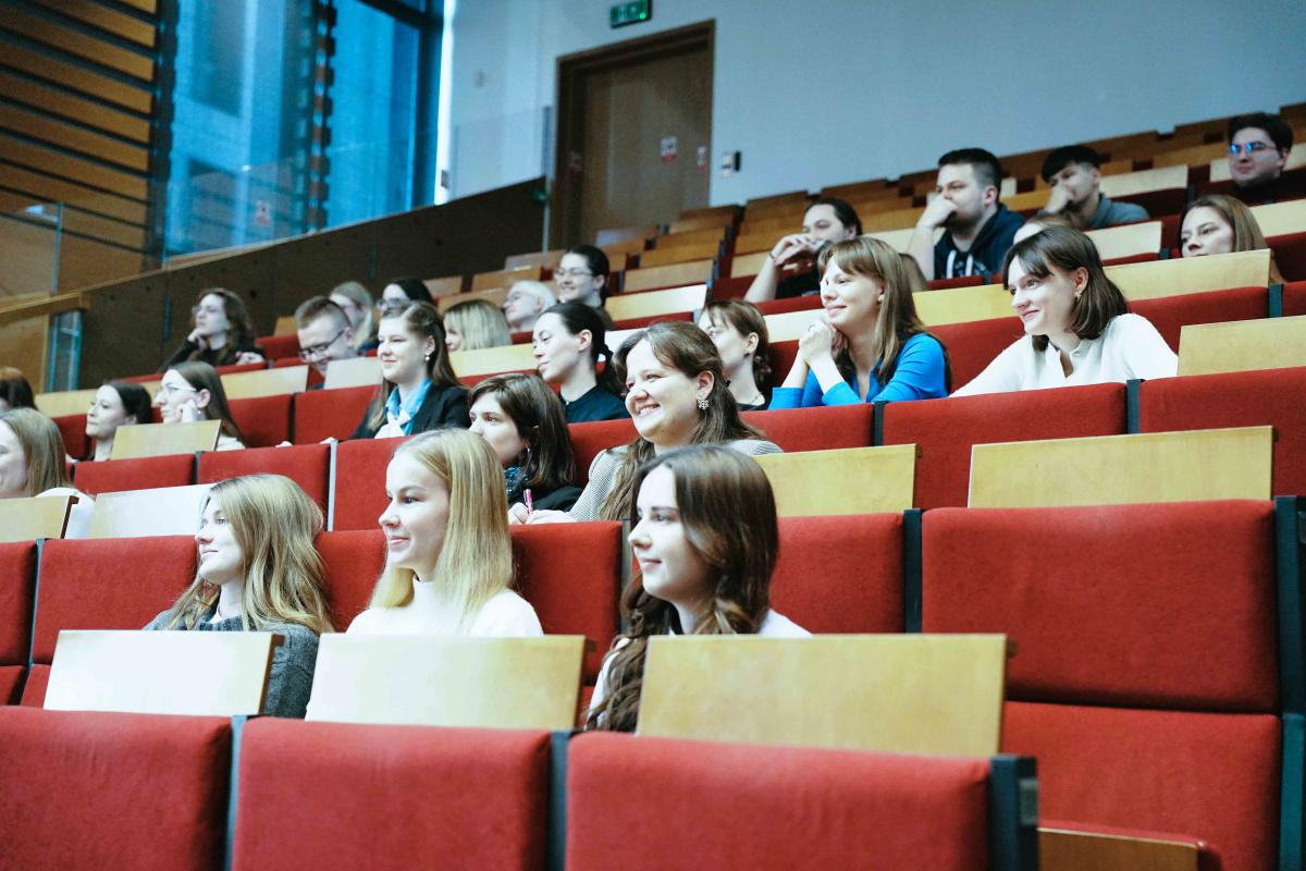 grupa ok. 30 kobiet i mężczyzn siedzi na auli wykładowej na czerwonych krzesłach. W tle, po lewej stronie widać duże okno i drzwi. 