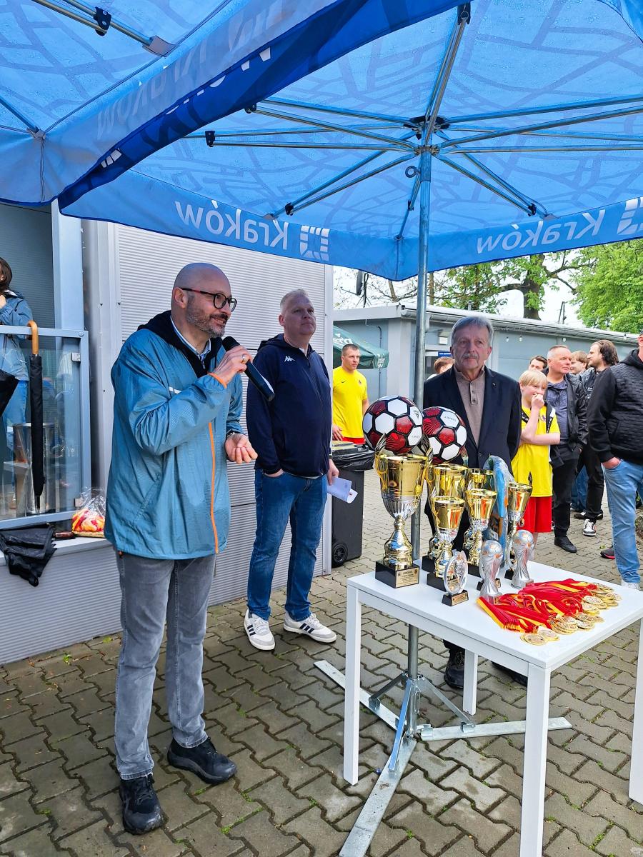Na zdjęciu znajduje się mężczyzna w szarych spodniach i błękitnej, sportowej kurtce, który przemawia do mikrofonu. Mężczyzna stoi obok białego stołu z pucharami i medalami. Na stole są złote puchary, na dwóch pucharach są położone piłki w kolorze czerwono-białym. Przed pucharami na stole leżą medale. Obok mężczyzny stoi dwóch mężczyzn w ciemnych ubraniach. Nad mężczyznami i stolikiem jest błękitny parasol z białym logo Krakowa i napisem Kraków. Mężczyźni stoją na nawierzchni z kostki brukowej, w tle jest grupa mężczyzn.