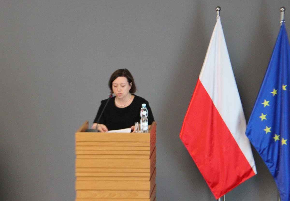 Kobieta stoi przy mównicy. Po prawej stronie znajdują się dwie flagi: Polski i Unii Europejskiej. 