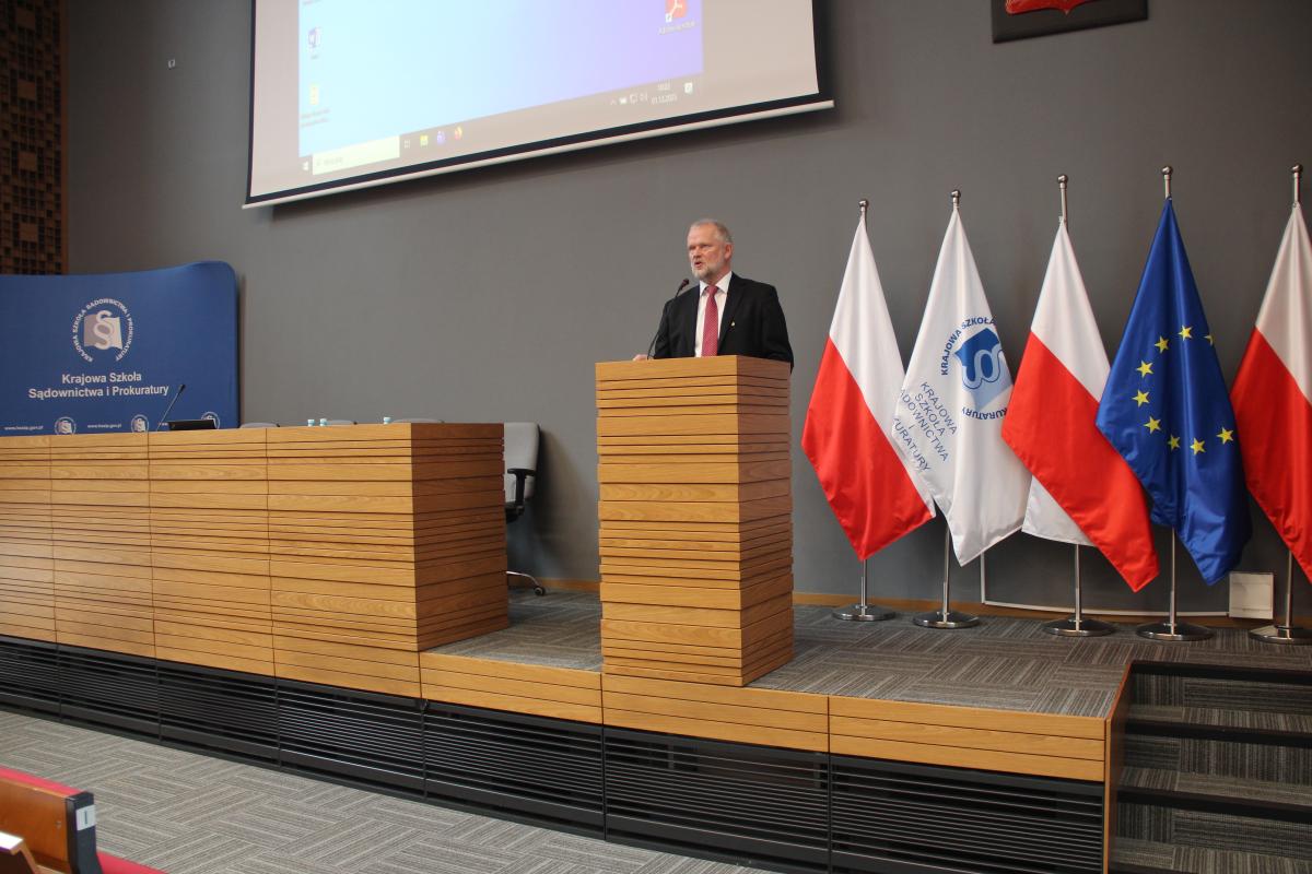 Zdjęcie przedstawia część auli wykładowej. Na pierwszym planie, za mównicą, widoczny jest mężczyzna  w ciemnym garniturze, białej koszuli i bordowym krawacie. Po prawej stronie zdjęcia znajdują się, ustawione w rzędzie, flagi: Polski, Unii Europejskiej oraz Krajowej Szkoły. W oddali, po lewej stronie, za katedrą, znajduje się baner w kolorze granatowym z napisem Krajowa Szkoła Sądownictwa i Prokuratury oraz logiem Krajowej Szkoły.