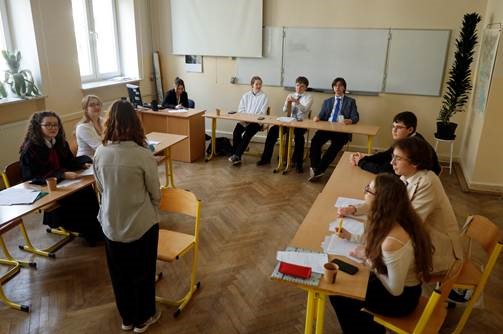Klasa. Ludzie siedzą przy stołach ustawionych w kształt litery U. Jedna osoba stoi na środku, wypowiada się. Na stołach są papiery, zeszyty i kubki. W klasie są duże okna, tablica i ekran projektora. Parkiet jest drewniany, na parapecie i w roku sali są kwiaty.