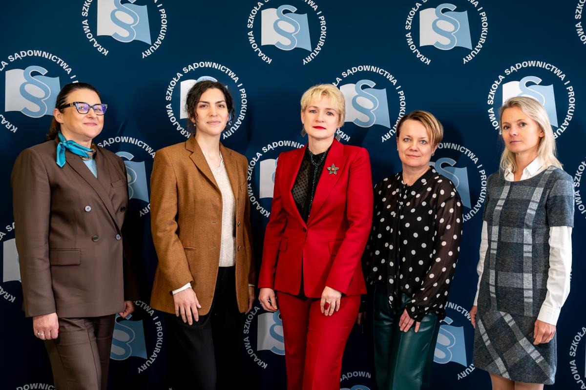 Na tle granatowej ścianki z logo Krajowej Szkoły Sadownictwa i Prokuratury stoi 5 kobiet. Od lewej Prokurator Aleksandra Wójcik -Zastępca Dyrektora Krajowej Szkoły, ubrana w garnitur  koloru czekoladowego, z błękitną apaszką na szyi, Agnieszka Matejczuk z Biura Rzecznika Praw Dziecka, ubrana w brązową marynarkę, sędzia Agnieszka Orłowska – Kierownik Działu Szkoleń Krajowej Szkoły, ubrana w czerwony garnitur z broszka w lewej klapie, kpt. SG Małgorzata -Komenda Główna Straży Granicznej, ubrana w bluzkę koloru czarnego w duże, białe kropki,  sędzia Beata Majewska z Działu Szkoleń Krajowej Szkoły, ubrana w sukienkę w kratę. 