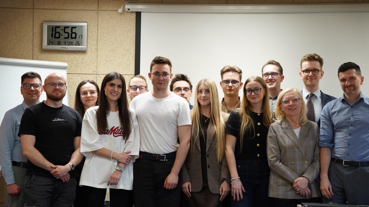 Zdjęcie przedstawia grupę uśmiechniętych uczestników zajęć, na tle białej tablicy oraz ściany, na której wisi zegar wyświetlający godzinę 16:56.
