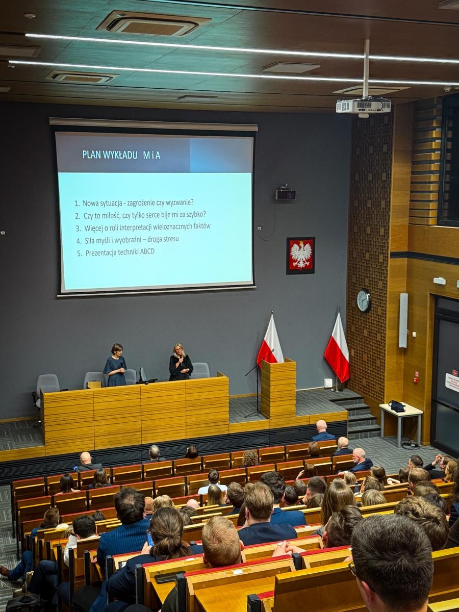 Zdjęcie przedstawia wykład dla aplikantów XVII rocznika aplikacji prokuratorskiej odbywający w auli KSSiP. Kilkudziesięciu uczestników siedzi na widowni, twarzami są zwróceni w stronę sceny. Na scenie, na podwyższeniu, przy mównicy stoją dwie kobiety, twarzami zwrócone w stronę audytorium. Za plecami kobiet znajduje się ekran powitalny, godło państwa oraz dwie flagi Polski. Na ekranie widoczny jest slajd z napisem „Plan wykładu M i A. 1. Nowa sytuacja -zagrożenie czy wyzwanie? 2. Czy to miłość, czy tylko serce bije mi za szybko? 3. Więcej o roli interpretacji wieloznacznych faktów. 4. Siła myśli i wyobraźni – droga stresu. 5. Prezentacja techniki ABCD