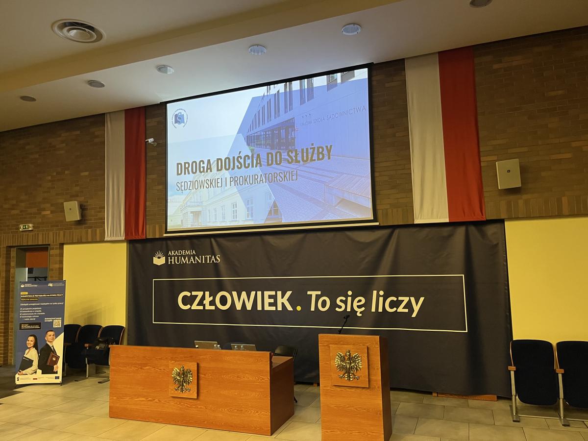 widok sali z dużym rzutnikiem, na  którym wyświetlony jest napis: Droga dojścia do służby sędziowskiej i prokuratorskiej. Poniżej znajduje się napis Akademia Humanitas. Człowiek. To się liczy. Z przodu stoi długi drewniany stół oraz miejsce do przemawiania, na których umieszczone jest godło Polski w złotym kolorze.