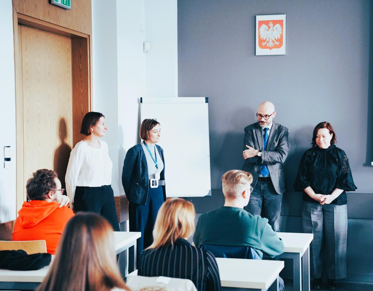 dwie kobiety i dwóch mężczyzn siedzi przy stolikach, plecami do obiektywu aparatu. Patrzą oni na trzy stojące kobiety i jednego mężczyznę zwróconych twarzami w ich stronę i stronę obiektywu aparatu. Na wprost na ścianie wisi godło Polski, w rogu, po lewej stronie stoi biała tablica. 