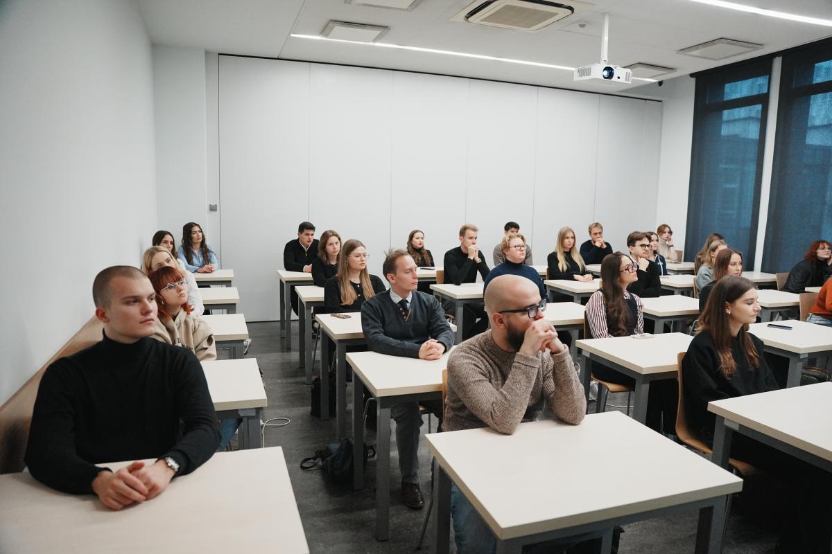 grupa około dwudziestu siedmiu kobiet i mężczyzn siedzi przy stolikach. Za nimi znajduje się duża biała ściana, a obok - po prawej stronie są duże okna zasłonięte roletami.