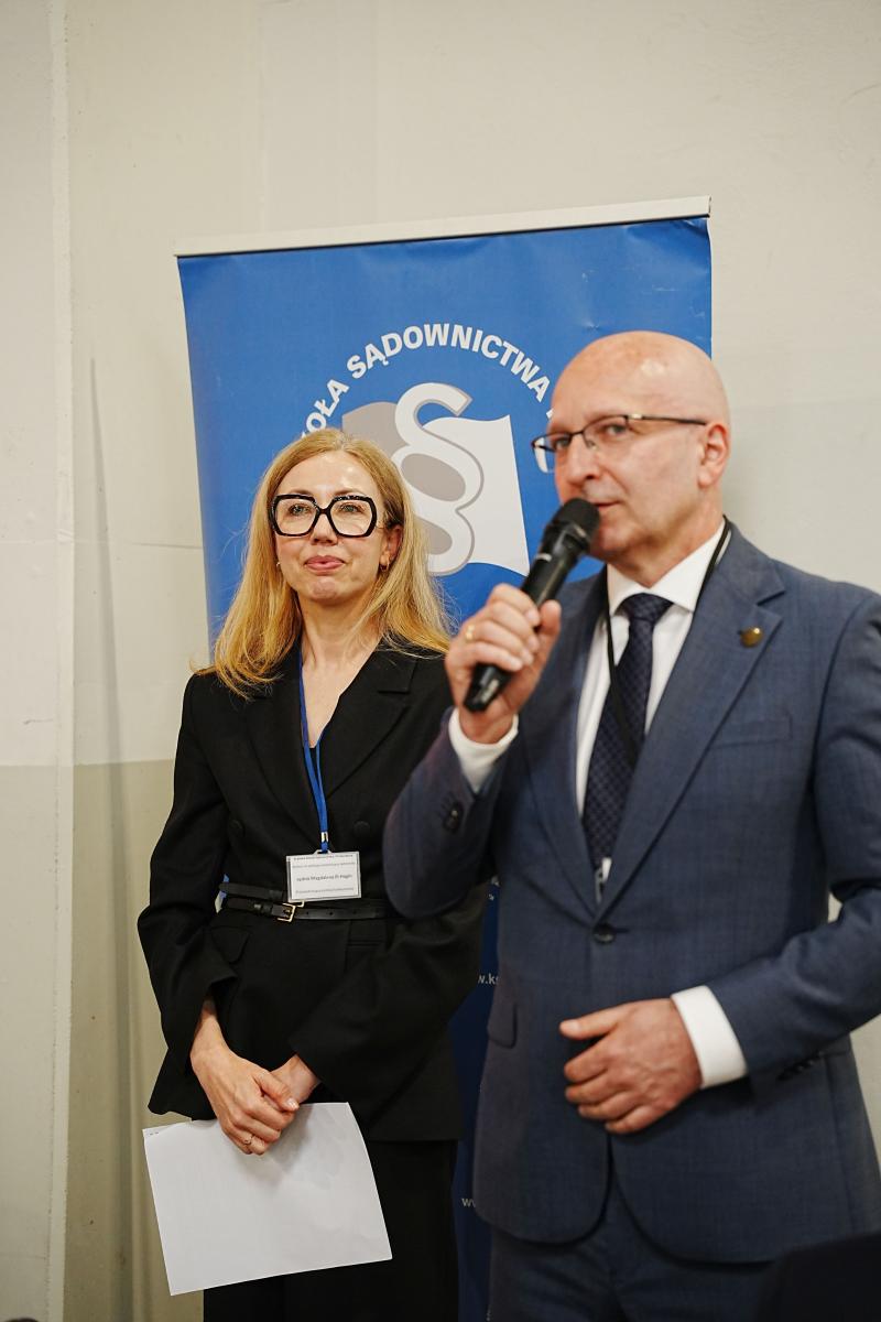 we wnętrzu pomieszczenia stoją dwie osoby w okularach, kobieta i mężczyzna. Mężczyzna jest ubrany w garnitur, białą koszulę i krawat. Stoi po prawej stronie i mówi do mikrofonu. Kobieta uśmiecha się. Jest ubrana w czarny żakiet i trzyma w opuszczonych rękach kartkę. W tle jest widoczny fragment banneru z logo.