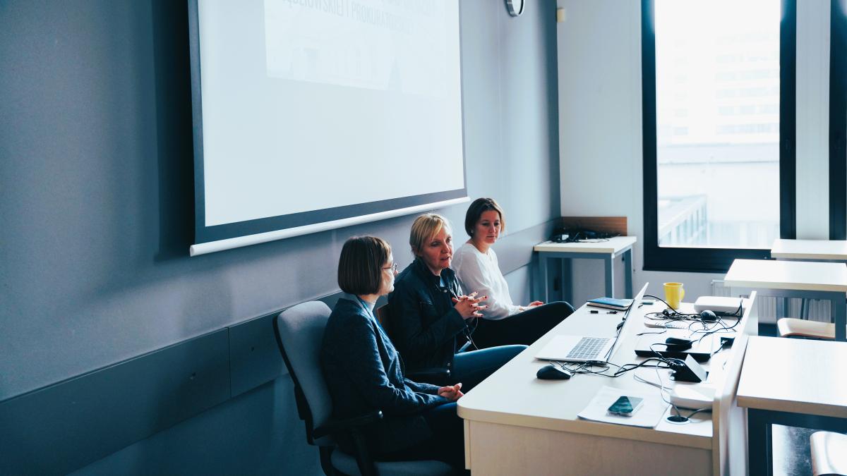 trzy kobiety siedzą przy biurku. Na biurku stoi laptop. Nad kobietami wisi tablica do wyświetlania prezentacji. Na ścianie obok znajdują się duże okna.