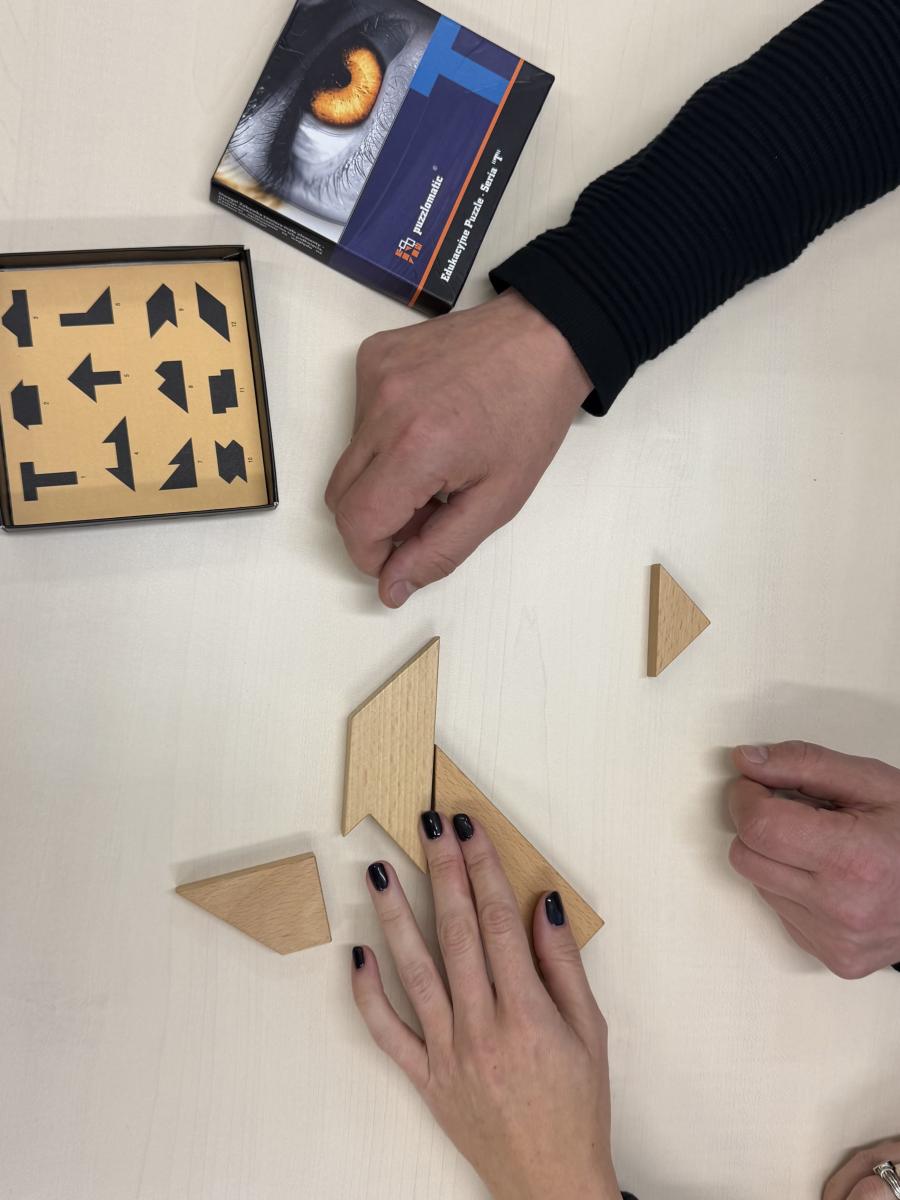 Na stoliku lewa dłoń damska z pomalowanymi na ciemny kolor paznokciami, położona na dwóch elementach drewnianych puzzli, które były układane podczas zajęć. Obok dwie męskie dłonie, oraz w górnym lewym rogu pudełko puzzli wraz z instrukcją.